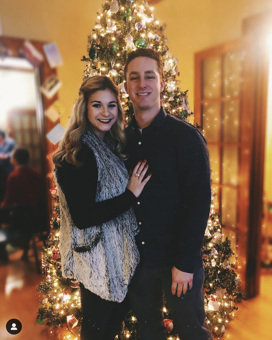 My boyfriend and I standing in front of a Christmas tree. The tree has lights and ornaments on it. We are both smiling, embracing, and looking at the camera.